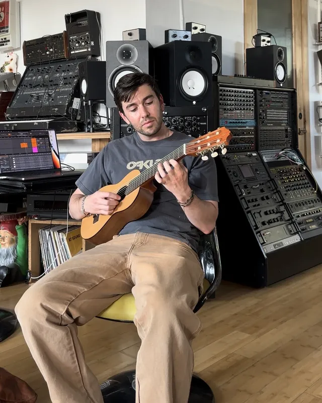 Tyler playing a small classical guitar in a studio full of synthesizers and speakers